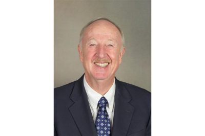 A man with gray hair and a mustache smiles for a headshot photo. He is wearing a navy blue suit, white dress shirt, and blue tie.