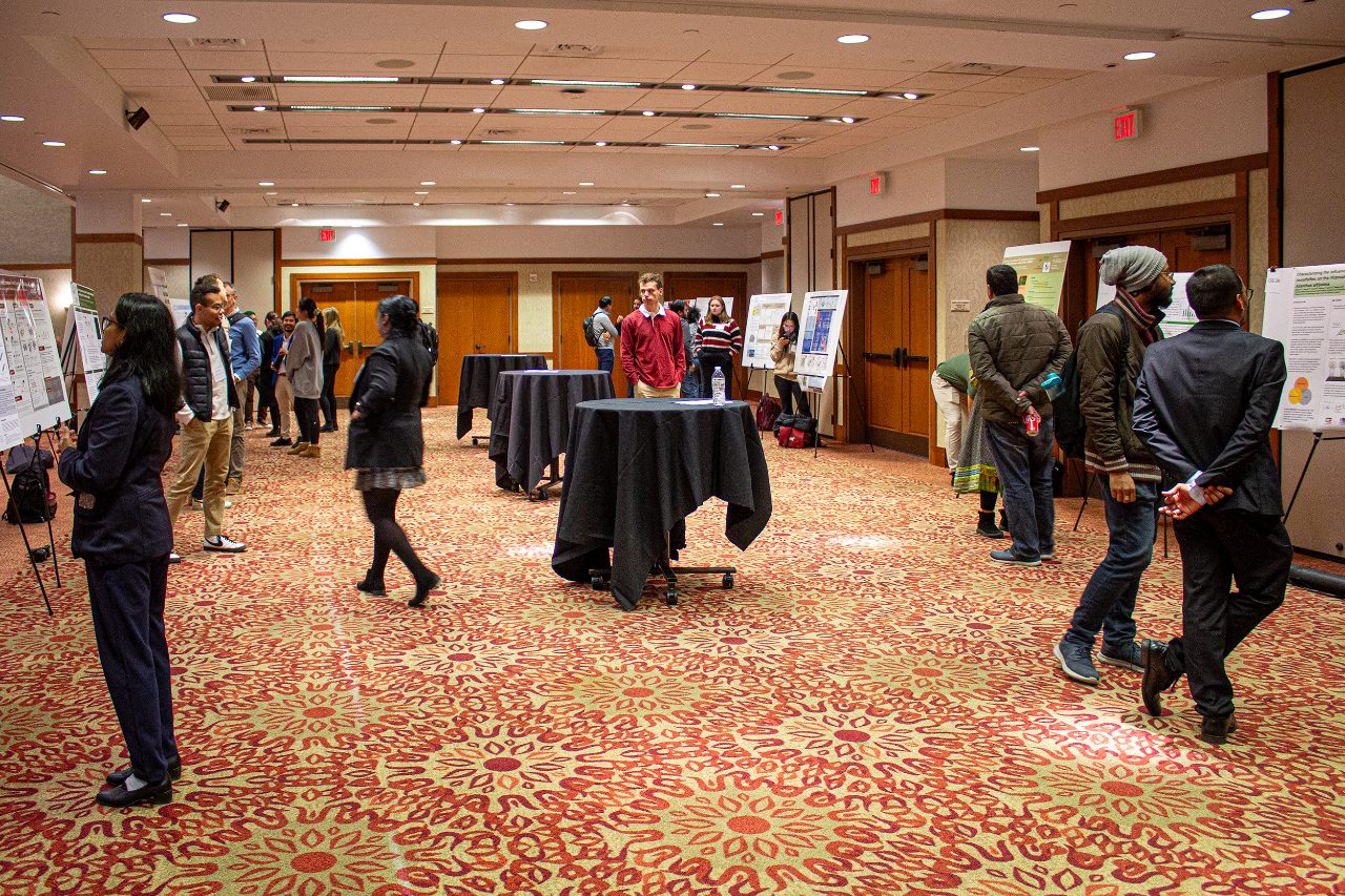 This shows a conference room with student posters lined up at the edges with attendees looking at the posters.